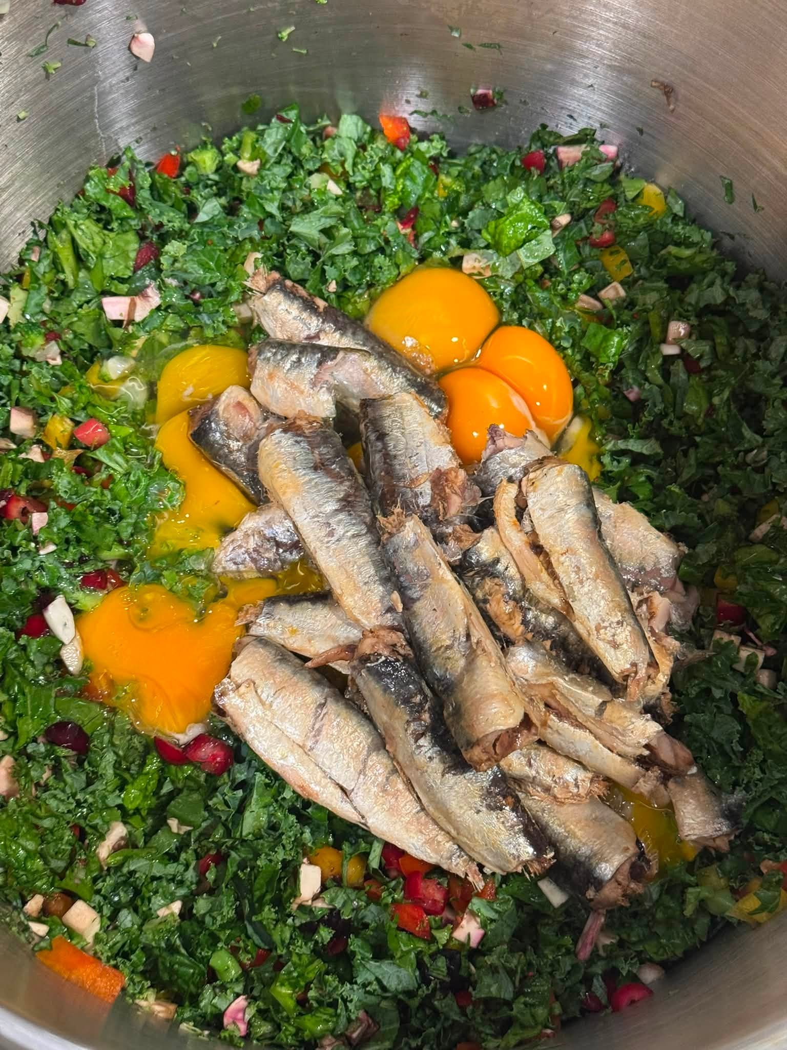 Sardines and cracked eggs over chopped greens and vegetables in a mixing bowl